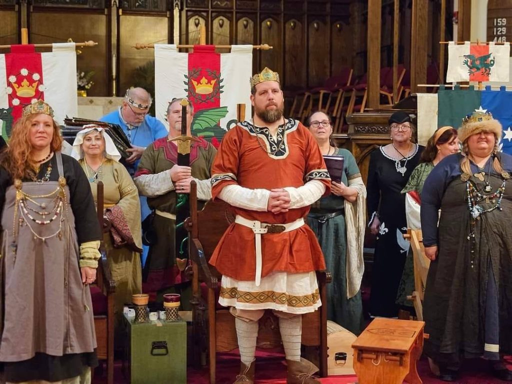 The king and queen of the Midrealm stand up in assembly with the Baron and Baroness of Ayreton with their court.